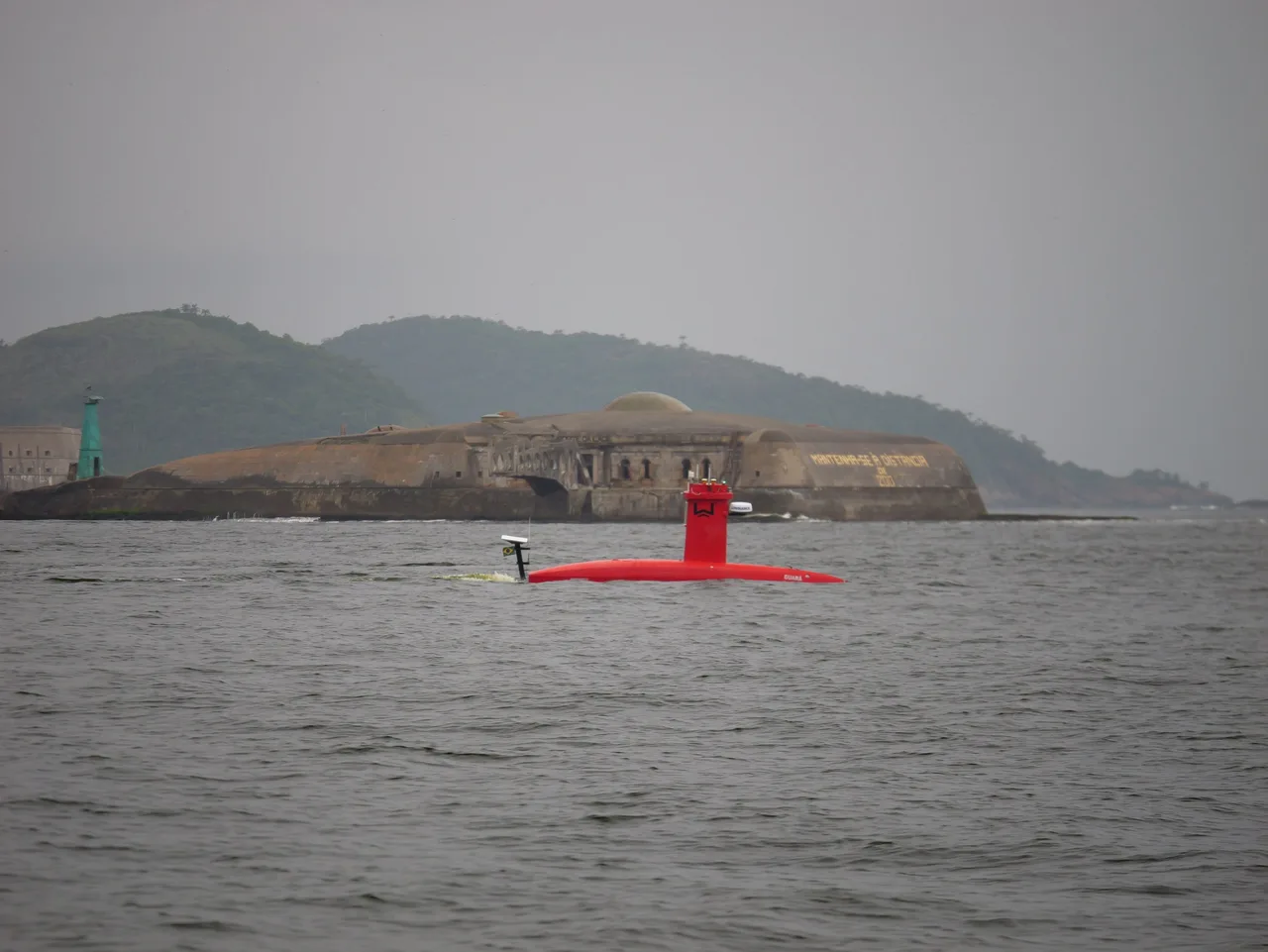 USV Exail DriX em navegação na Baía de Guanabara, com um forte e morros ao fundo.