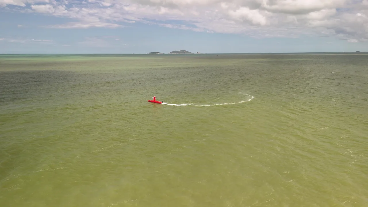 USV Exail DriX em linha de levantamento em mar aberto, visto do alto, com a costa ao fundo.