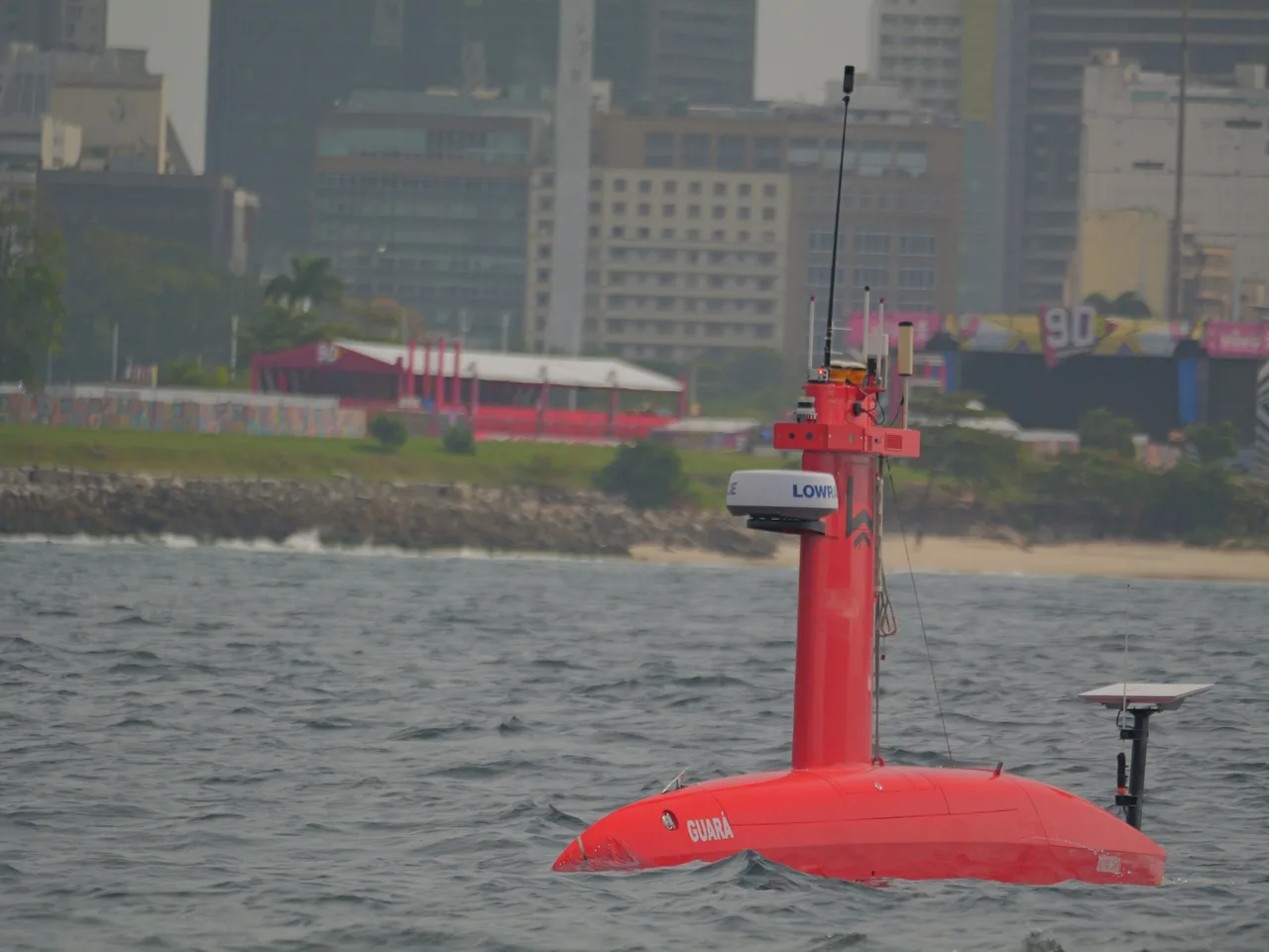 USV Exail DriX próximo à orla na Baía de Guanabara, mastro com antenas e casco vermelho.