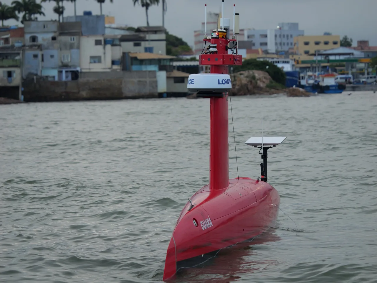 USV Exail DriX em águas abrigadas, visto de frente, com casas da orla ao fundo.