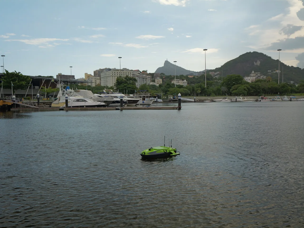 USV Hi-Target iBoat BS15 em navegação em uma marina, com barcos e morros ao fundo.