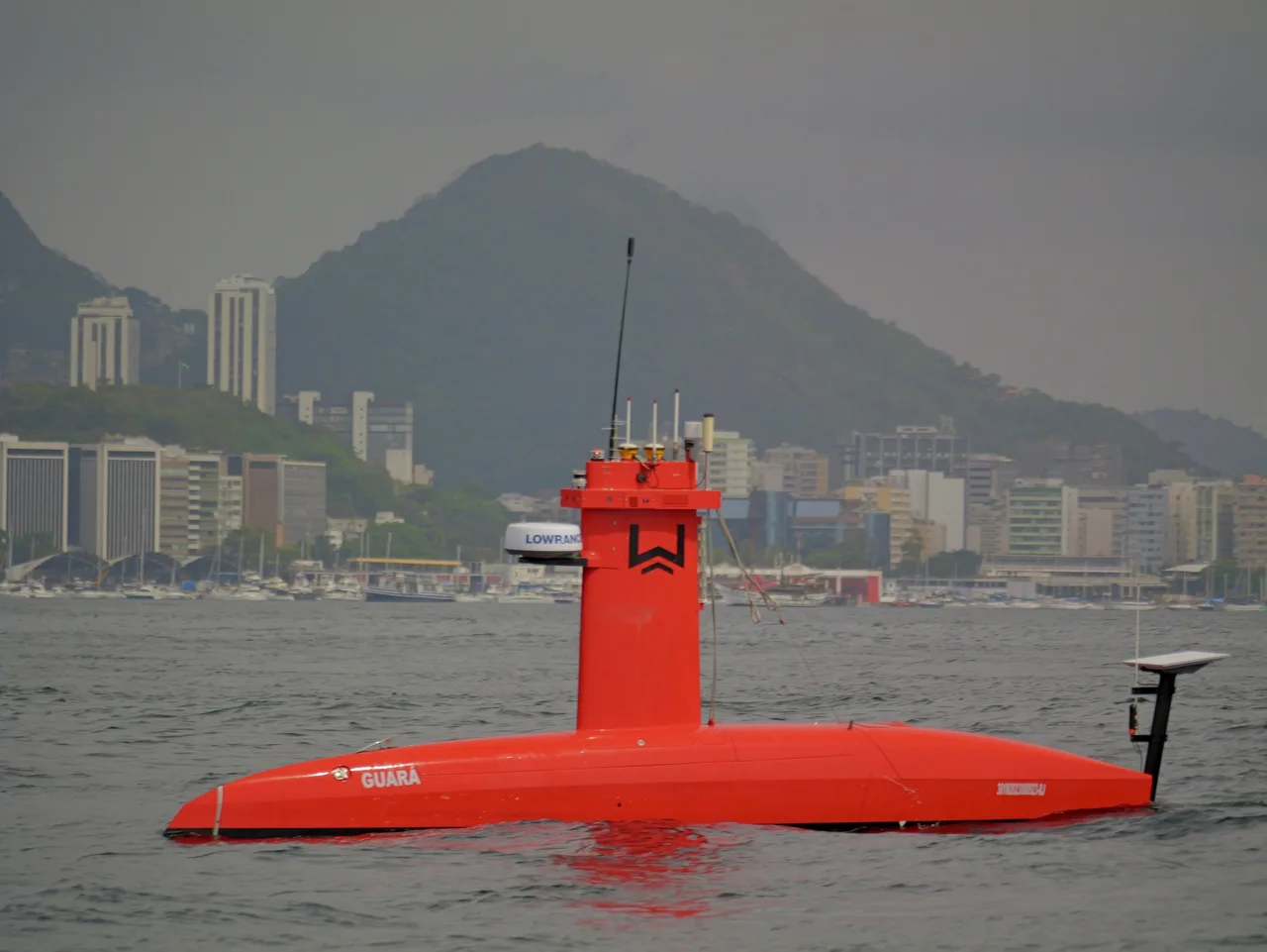 USV Exail DriX de perfil em navegação na Baía de Guanabara, com morro e prédios ao fundo.