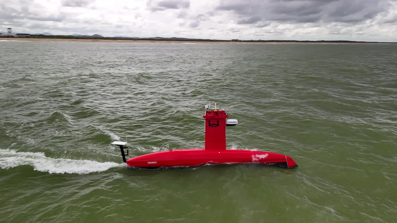 USV Exail DriX em navegação em mar aberto, visto de lado pelo drone, com a esteira na água.