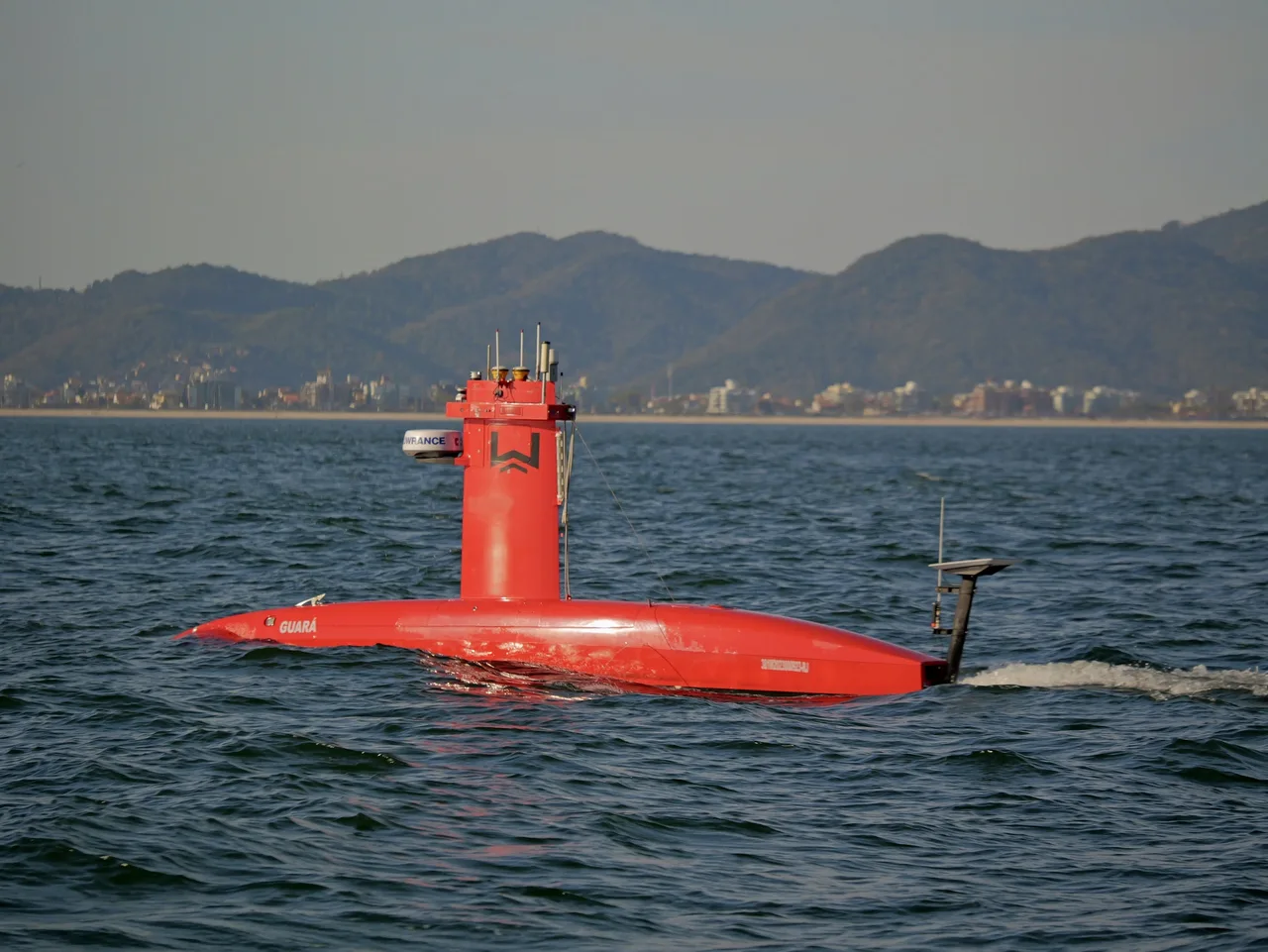 USV Exail DriX em navegação, casco vermelho e mastro com antenas, morros ao fundo.