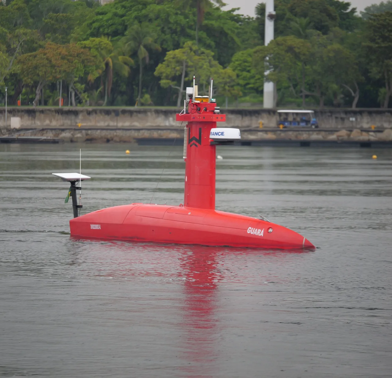 USV Exail DriX em navegação na Baía de Guanabara, com o reflexo do casco na água e árvores ao fundo.