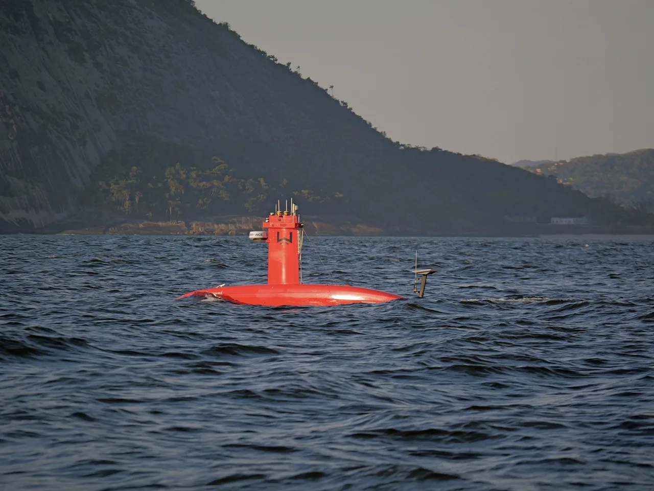 USV Exail DriX navegando próximo à costa, com encosta de vegetação ao fundo.